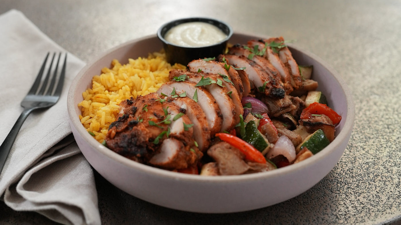 A bowl of prepared food on a round table, featuring sliced grilled chicken with yellow rice, mixed vegetables, and a small side of white sauce. The bowl is placed on a folded napkin with a fork, in a softly blurred indoor setting.